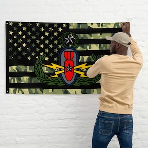 以下が含まれることがあります： 中央に黒と黄色のEOD（爆発物処理）バッジが付いた迷彩柄のアメリカ国旗。旗は、ベージュのシャツと青いジーンズを着用した人が白いレンガの壁に掛けています。