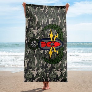 以下が含まれることがあります： 迷彩柄のビーチタオルで、海軍のEOD（爆発物処理）の徽章が描かれています。徽章は、稲妻と月桂樹の輪で飾られた爆弾です。