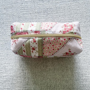 May include: A rectangular, patchwork-style zippered pouch. The pouch features a variety of floral patterns in pink, red, and green on a light background. The zipper is gold-colored, and a small red bow is attached to one end.