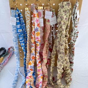 May include: A display of fabric keychains with floral patterns in various colors, including blue, yellow, pink, and white. The keychains are hanging from a wooden pegboard with gold-colored hardware. Each keychain has a small tag that reads "Lincoln & Co."