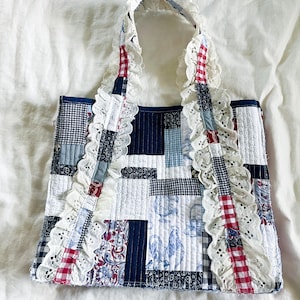 May include: A patchwork tote bag with a quilted design. The bag features a variety of fabric patterns in blue, white, and red, including gingham, floral, and solid colours. The handles are adorned with white lace trim.