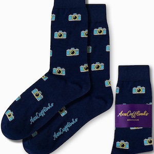 Op de afbeelding: Een paar donkerblauwe sokken met een herhalend patroon van blauwe en gele camera's. De sokken zijn gevouwen en worden getoond met een verpakkingsetiket met de tekst "AusCufflinks AUSTRALIA".