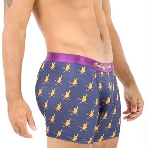 May include: Navy blue boxer shorts featuring a repeating pattern of yellow skiers. The purple waistband has the text "Aus Cufflinks" in gold. The shorts are close-fitting.