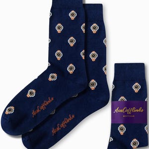Könnte beinhalten: Marineblaue Socken mit einem sich wiederholenden Muster aus goldenen und weißen Uhrendesigns. Die Socken tragen den Text "AusCufflinks" in kursiver Schrift. Ein Paar Socken wird mit einer gefalteten und mit einem lila Band umwickelten Socke gezeigt.
