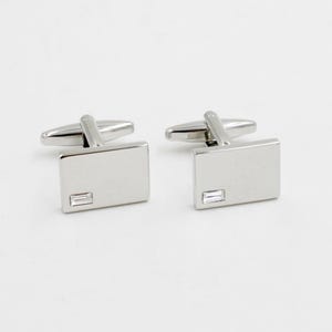 May include: A pair of silver cufflinks with a rectangular design. Each cufflink has a small, rectangular, clear stone. The cufflinks have a shiny, polished finish and a classic, elegant style.