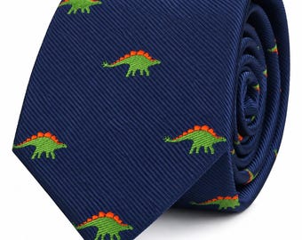 May include: A navy blue necktie featuring a repeating pattern of small, embroidered green stegosaurus dinosaurs with bright orange plates along their backs. The fabric has a subtle diagonal ribbed texture throughout the entire surface.