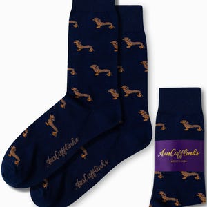 May include: Navy blue socks featuring a repeating pattern of brown dachshund dogs. The socks are branded with "AusCufflinks" in gold script. A pair of socks is shown next to a single sock with a purple band.