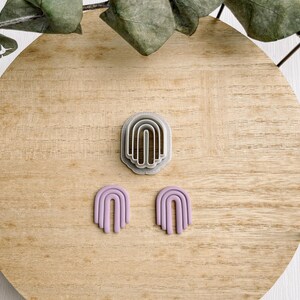 May include: A set of two purple polymer clay earrings shaped like arches. The earrings are displayed on a wooden surface with a silver arch-shaped cookie cutter.