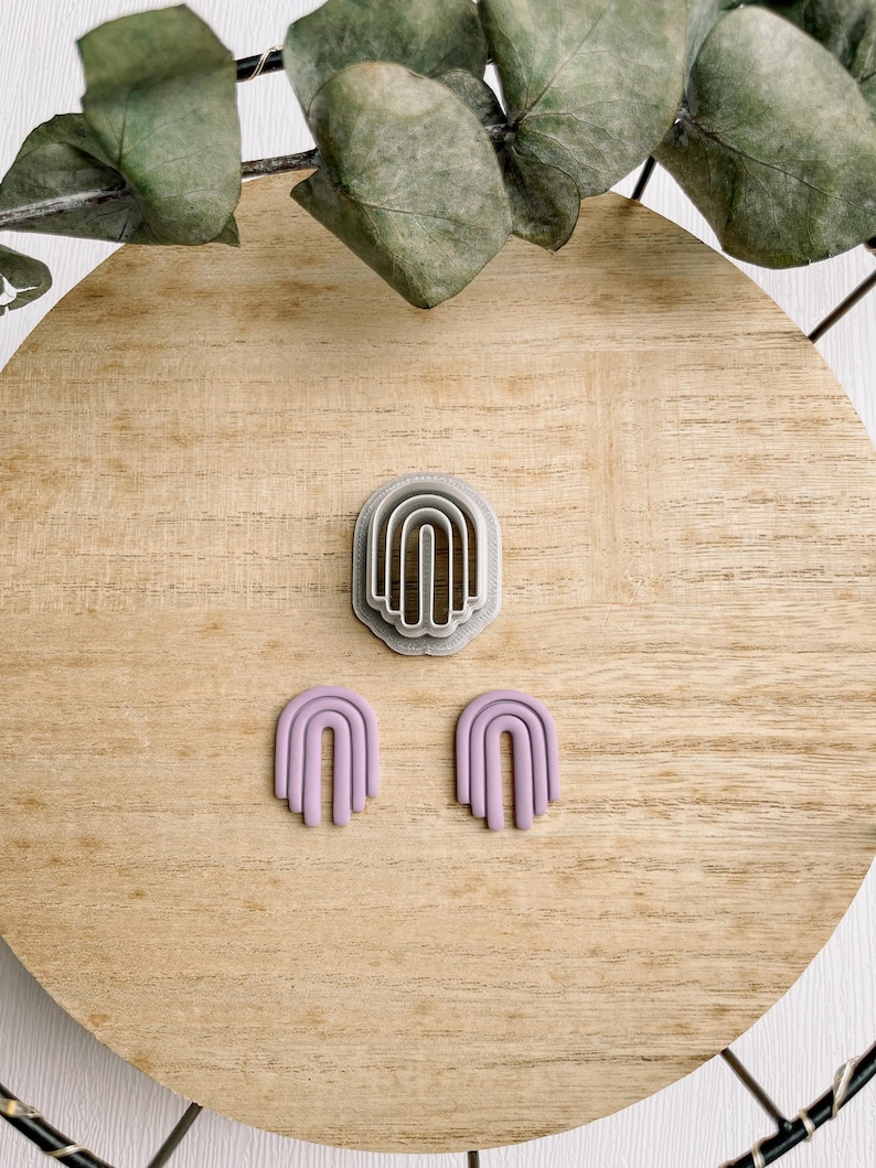 May include: A set of two purple polymer clay earrings shaped like arches. The earrings are displayed on a wooden surface with a silver arch-shaped cookie cutter.