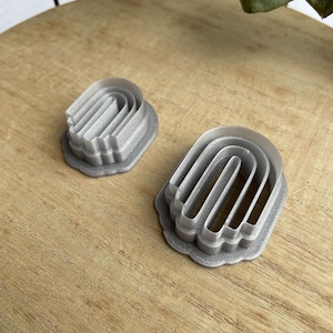 May include: Two silver metal cookie cutters shaped like arches. The cutters are on a wooden surface.