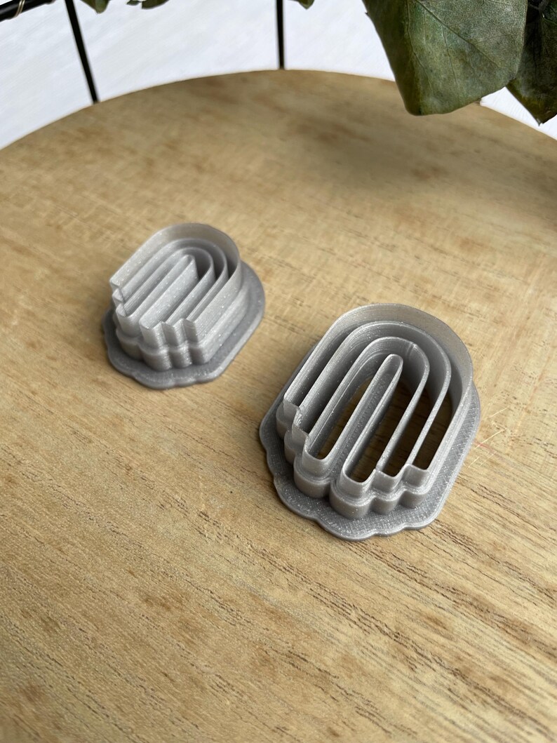 May include: Two silver metal cookie cutters shaped like arches. The cutters are on a wooden surface.