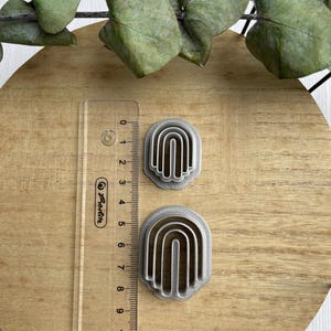 May include: Two silver-colored metal cookie cutters shaped like rainbows. The cutters are on a wooden surface with a ruler next to them.