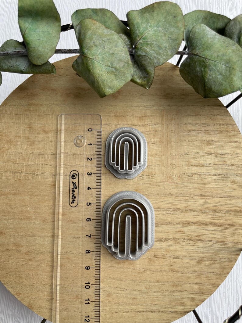 May include: Two silver-colored metal cookie cutters shaped like rainbows. The cutters are on a wooden surface with a ruler next to them.