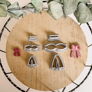 Pode incluir: Um conjunto de seis cortadores de biscoitos de plástico cinza em forma de laço, vestido e sapatos. Também são mostrados dois laços rosa sobre uma superfície de madeira.