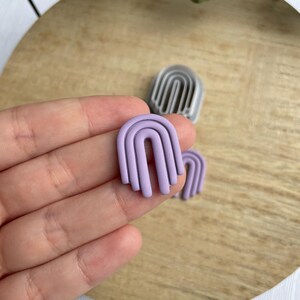 May include: A pair of lavender polymer clay earrings shaped like arches. The earrings are layered to create a three-dimensional effect.