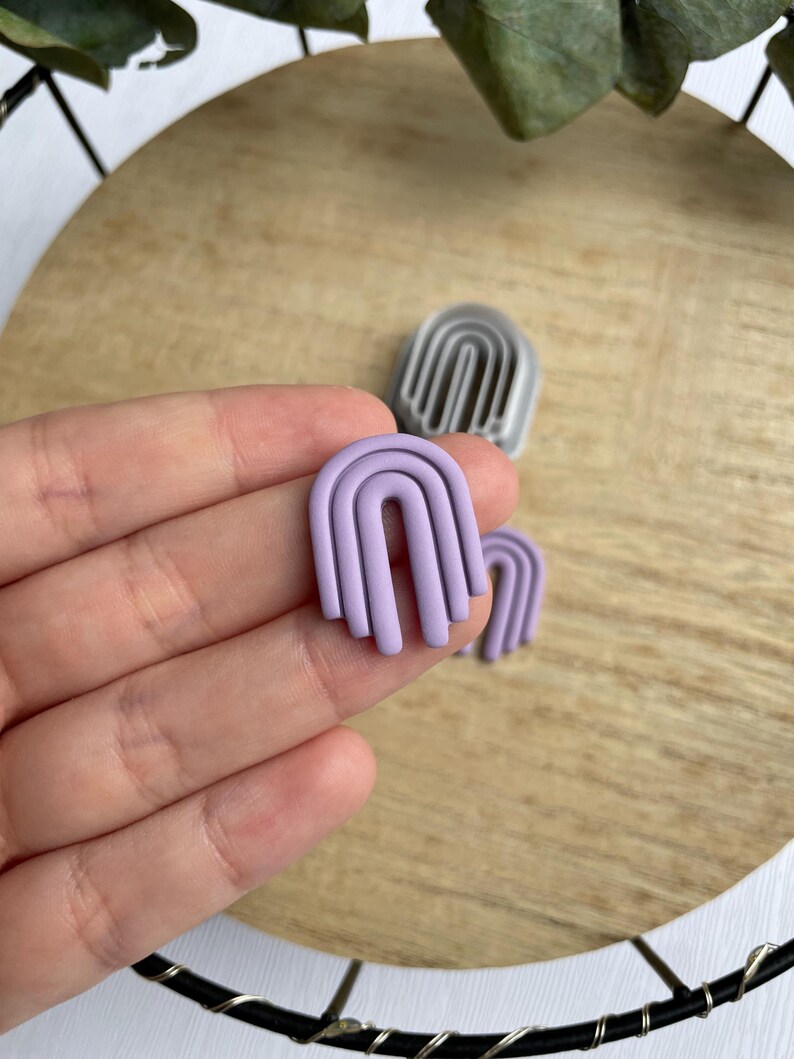 May include: A pair of lavender polymer clay earrings shaped like arches. The earrings are layered to create a three-dimensional effect.
