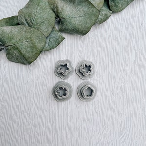 Puede incluir: Cuatro cortadores de galletas de plástico gris en forma de flor, estrella, flor con un círculo central y flor con un círculo central y un anillo alrededor. Los cortadores están sobre una superficie de madera blanca.