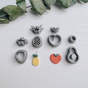 Könnte beinhalten: Ein Set mit 10 grauen Plastik-Ausstechern in Form von Ananas, Herz, Avocado, Pfirsich und anderen Fruchtformen. Die Ausstecher sind auf einer weißen Oberfläche mit einem Zweig grüner Eukalyptusblätter angeordnet.