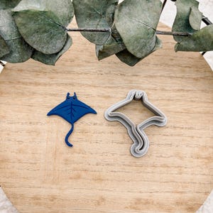 May include: Two stingray-shaped items on a wooden surface. One is a vibrant blue, the other a grey cookie cutter. The blue stingray has a detailed design. The items are arranged on a light brown wooden surface, with eucalyptus leaves in the background.