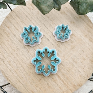 Peut inclure: Trois emporte-pièces bleus en forme de flocon de neige sur une surface en bois. Les emporte-pièces sont de différentes tailles, avec un intérieur bleu clair et un bord blanc. Conçu pour la pâtisserie et les projets créatifs.