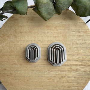 May include: Two silver cookie cutters shaped like arches, one larger than the other, on a wooden surface.