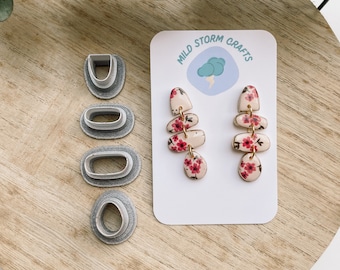 May include: A pair of floral earrings and four oval-shaped clay cutters are displayed on a wooden surface. The earrings feature a stacked design with a pink floral pattern. The cutters are gray and vary in size and shape.