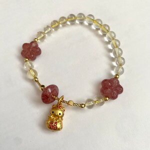 May include: A bracelet made with clear and pink beads, with a gold charm of a cat holding a gold coin with Chinese characters. The charm is attached to a pink bead.