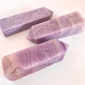 Puede incluir: Cuatro obeliscos de cristal de amatista con base rectangular y parte superior puntiaguda. Los cristales tienen un tono púrpura claro con variaciones naturales de color y textura. Los obeliscos están dispuestos sobre una superficie blanca.