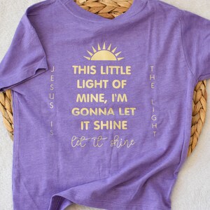 May include: Purple short-sleeved t-shirt with gold lettering that reads "This little light of mine, I'm gonna let it shine let it shine" and "Jesus is the light".