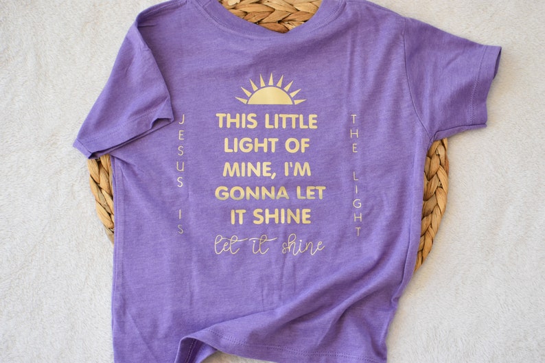 May include: Purple short-sleeved t-shirt with gold lettering that reads "This little light of mine, I'm gonna let it shine let it shine" and "Jesus is the light".