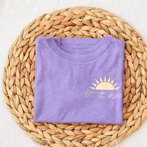 May include: A purple t-shirt with a gold sun graphic and the text "Let it shine".
