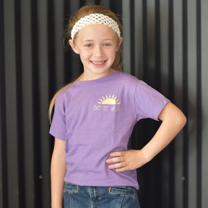 May include: A young person wearing a purple t-shirt with a gold sun graphic and the text "Let it shine". They are also wearing a white headband and a blue denim skirt.