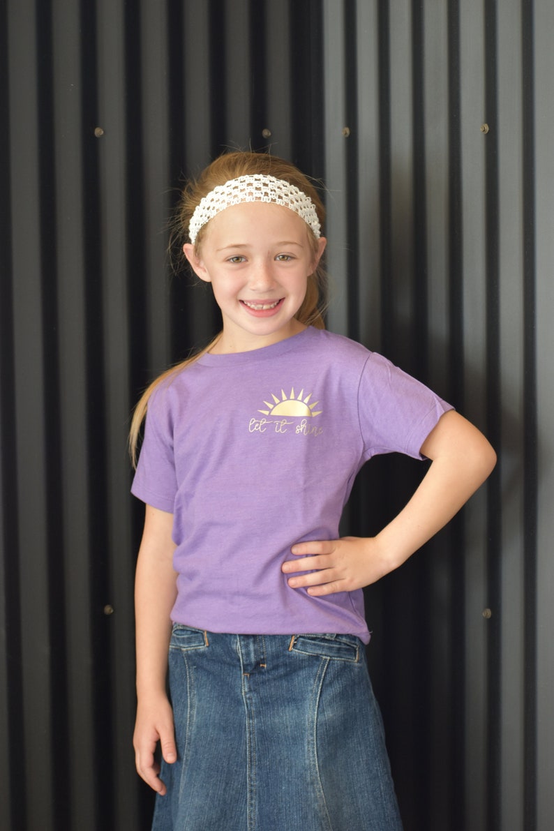 May include: A young person wearing a purple t-shirt with a gold sun graphic and the text "Let it shine". They are also wearing a white headband and a blue denim skirt.