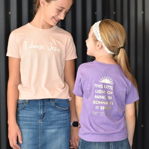 May include: Two girls wearing t-shirts with religious messages. The girl on the left is wearing a light pink t-shirt with the text "I choose Jesus" in white. The girl on the right is wearing a purple t-shirt with the text "This little light of mine, I'm gonna let it shine" in gold.