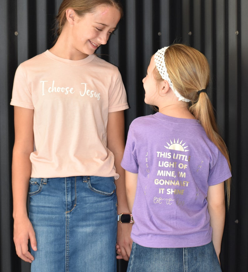 May include: Two girls wearing t-shirts with religious messages. The girl on the left is wearing a light pink t-shirt with the text "I choose Jesus" in white. The girl on the right is wearing a purple t-shirt with the text "This little light of mine, I'm gonna let it shine" in gold.