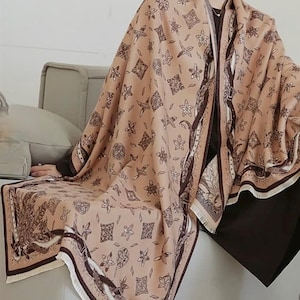 Puede incluir: Una bufanda de color marrón y beige con un diseño geométrico. La bufanda tiene un borde con flecos y está hecha de un material suave y cálido.
