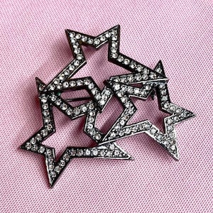 May include: A brooch featuring three interconnected star-shaped frames made of dark gunmetal-toned metal. Each star outline is densely set with small, clear, sparkling rhinestones that follow the geometric contours of the five-pointed shapes.
