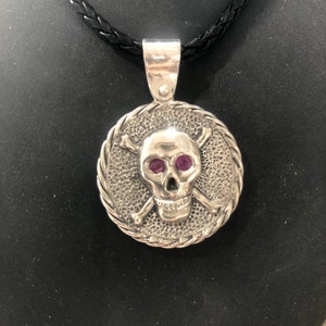 May include: A silver skull pendant with crossed bones and purple gemstone eyes, hanging from a black leather cord.