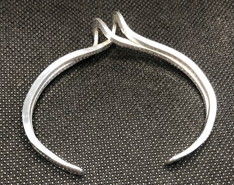 Könnte beinhalten: Ein silbernes Armreif-Armband mit einem einzigartigen, abstrakten Design. Das Armband ist offen und hat einen einfachen, minimalistischen Stil.