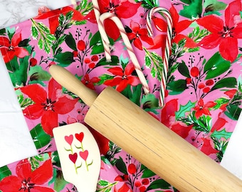 Könnte beinhalten: Festliche Küchenszene mit einem rosa Küchentuch mit Blumenmuster mit roten Weihnachtssternen und grünen Blättern. Zuckerstangen, eine hölzerne Teigrolle und ein Spatel mit Herzmotiven sind auf dem Tuch angeordnet und schaffen ein Urlaubs-Thema.