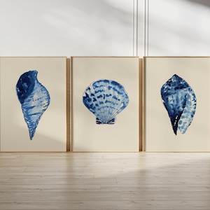 May include: Three framed art prints featuring watercolor illustrations of seashells in shades of blue. The prints are displayed on a light-colored wall and rest on a wooden floor. The frames have a gold finish.