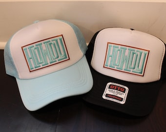 May include: Two trucker hats with a white front panel and a black mesh back. The front panel of both hats has a brown and white rectangular border with the word "HOWDY" in turquoise lettering. The hat on the left has a light blue front panel and the hat on the right has a black front panel with a white "OTTO" label.