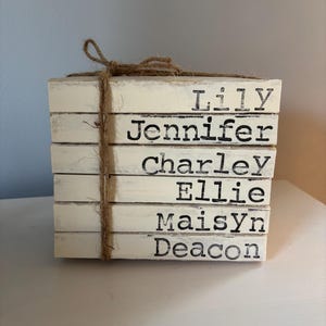 May include: A stack of six off-white wooden blocks tied with twine. Each block has a name printed in black: Lily, Jennifer, Charley, Ellie, Maisyn, and Deacon. The blocks are stacked vertically.