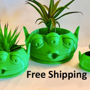 May include: Three green alien-shaped planters with three eyes each. The planters are made of 3D printed plastic and are filled with small green plants. The text "Free Shipping" is visible at the bottom of the image.