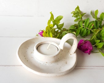 Op de afbeelding: Witte keramische kaarsenhouder met een kleine, ronde kaars in het midden. De houder heeft een uniek handvat en een gespikkelde afwerking.