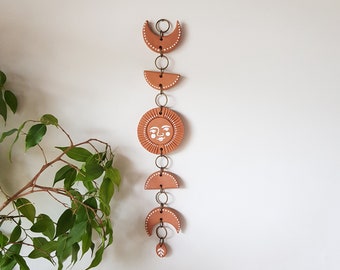 May include: Terracotta sun and moon wall hanging with white painted details. The sun has a face and the moons are crescent shapes. The pieces are connected by gold metal rings.
