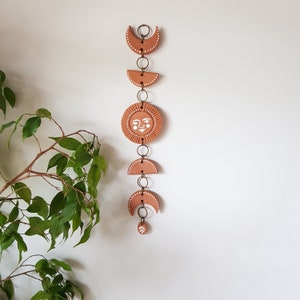 May include: Terracotta sun and moon wall hanging with white painted details. The sun has a face and the moons are crescent shapes. The pieces are connected by gold metal rings.