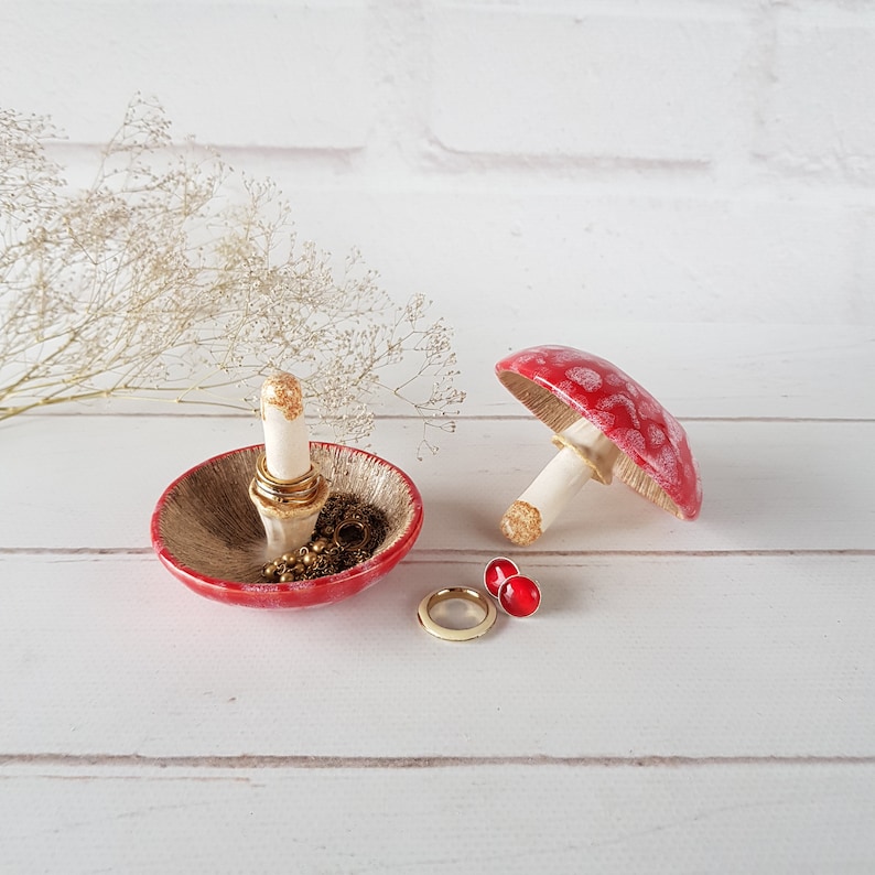 May include: A red and gold ceramic mushroom-shaped ring holder and a matching mushroom figurine. The ring holder has a gold interior and a white ring stand. A gold ring and red earrings are also present.