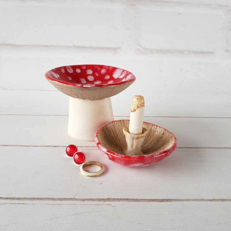 May include: Ceramic mushroom-shaped ring holders and a mushroom-shaped trinket dish. The ring holders have a red top with white spots and a beige underside. Two red stud earrings and a gold ring are also present.