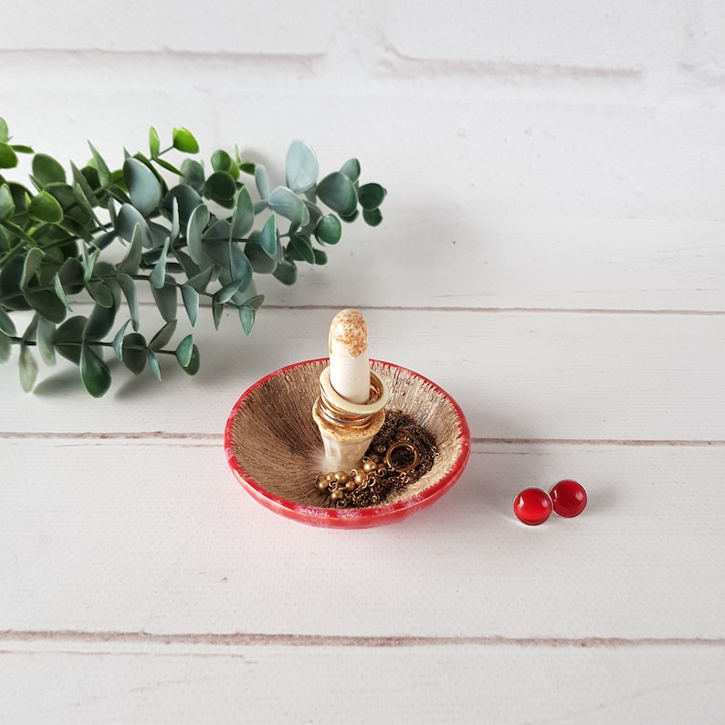 May include: A ceramic ring holder with a red rim and a beige interior. A central post holds rings, and the dish contains a gold necklace. Two red glass beads are on the side. The holder is on a white wooden surface.